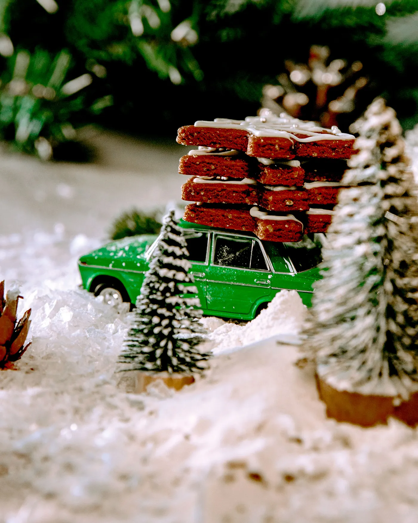 A green toy car carrying Christmas cookies through a snow-covered festive scene with trees.