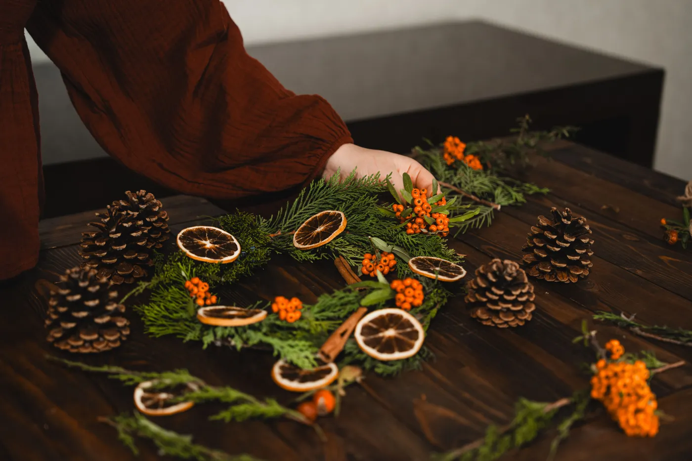 A person arranges a natural Christmas wreath with pinecones and dried orange slices on a wooden table.