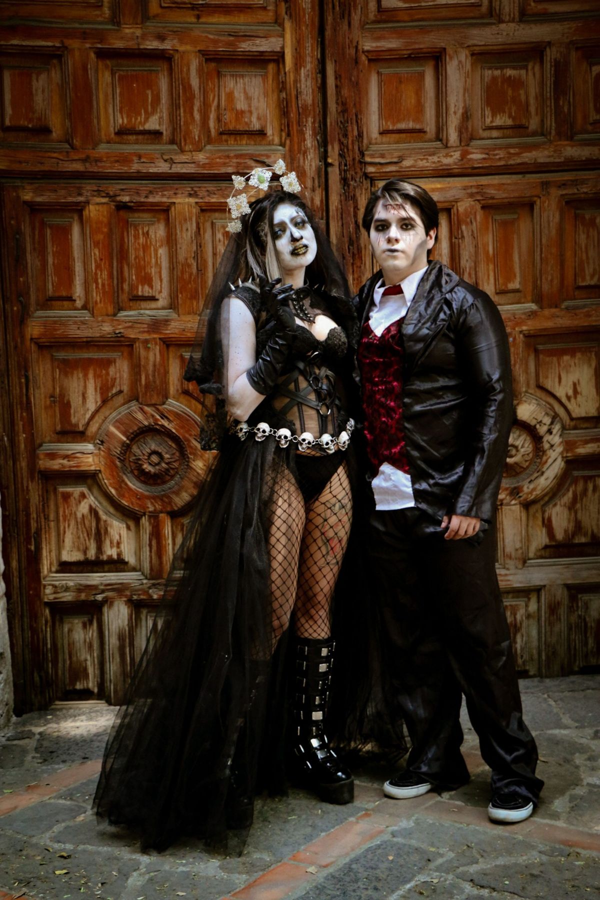 A spooky couple in Gothic Halloween costumes stands before a vintage wooden door.