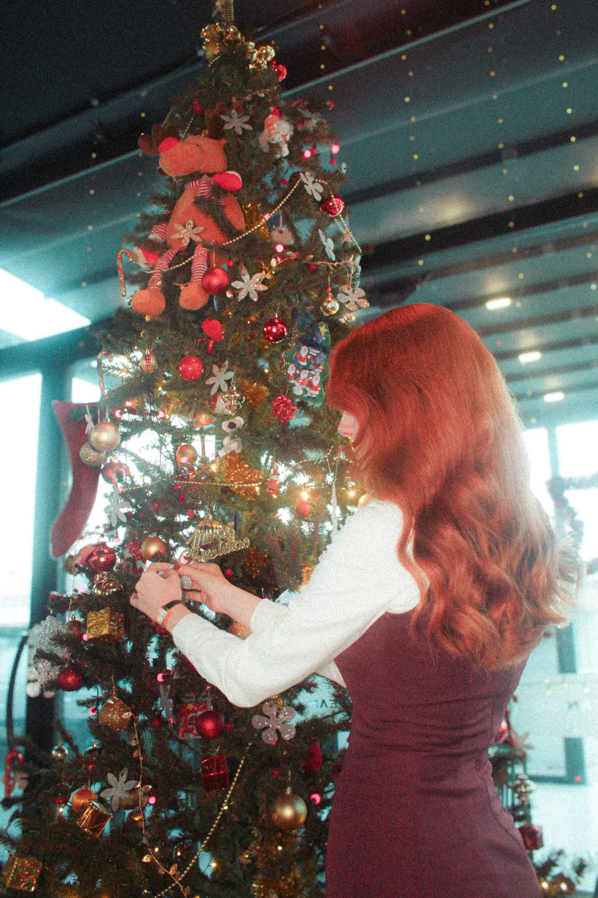 A woman decorates a Christmas tree indoors with festive ornaments and lights.