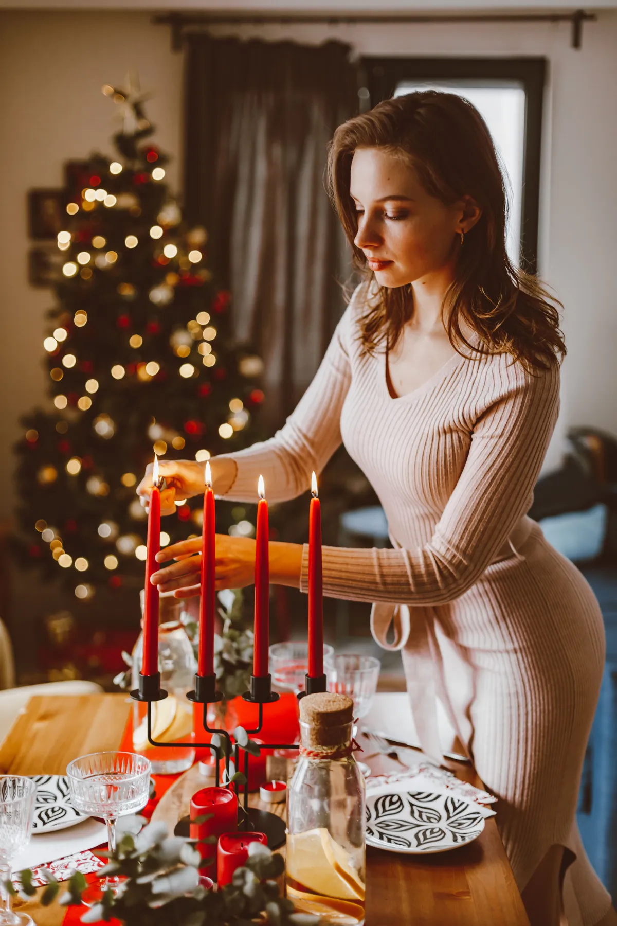A woman decorates a festive holiday table with lit candles in a cozy, Christmas-themed room.