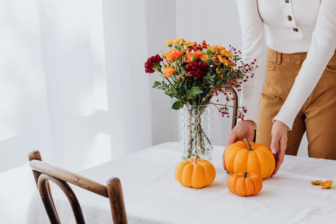 A woman decorates a table with pumpkins and a floral arrangement in a glass vase for fall.