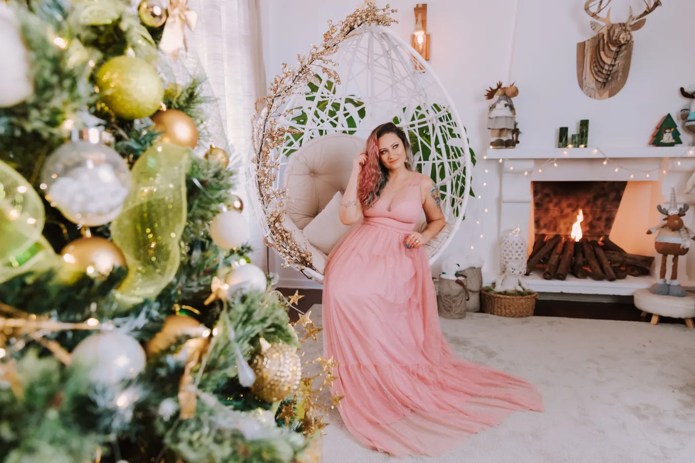 A woman in a pink dress sits elegantly in a hammock chair beside a decorated Christmas tree and fireplace.