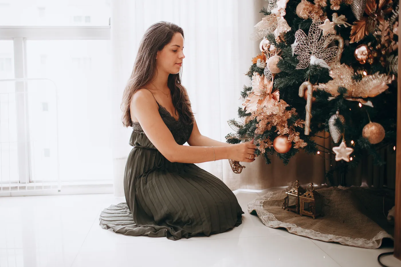 A woman in an elegant dress decorates a festive Christmas tree indoors, creating a warm holiday atmosphere.