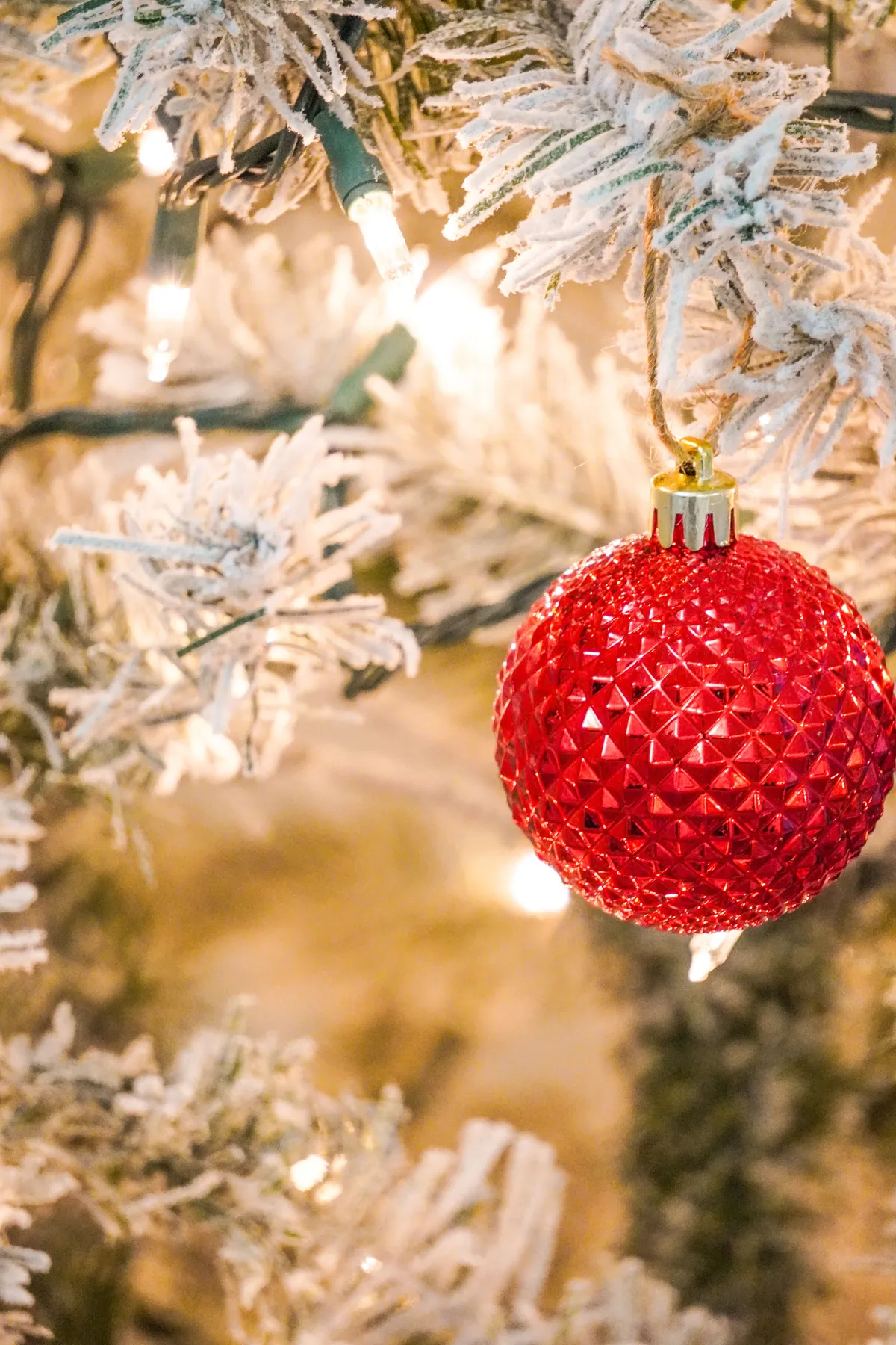 Close-up of a red ornament hanging on a frosted Christmas tree with warm glowing lights.