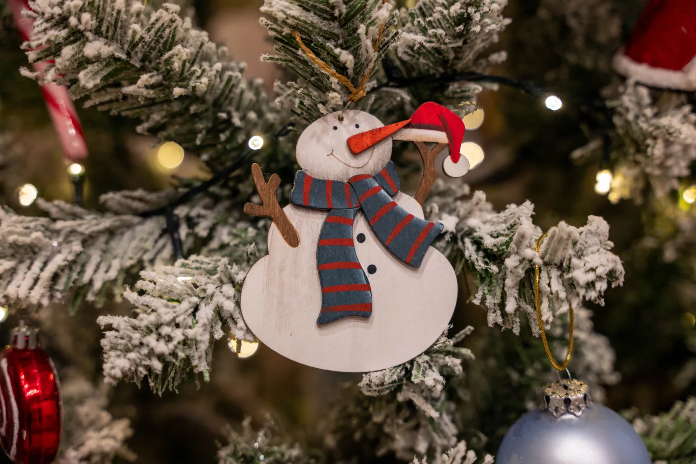 Close-up of a snowman ornament on a flocked Christmas tree, illuminated with holiday lights.