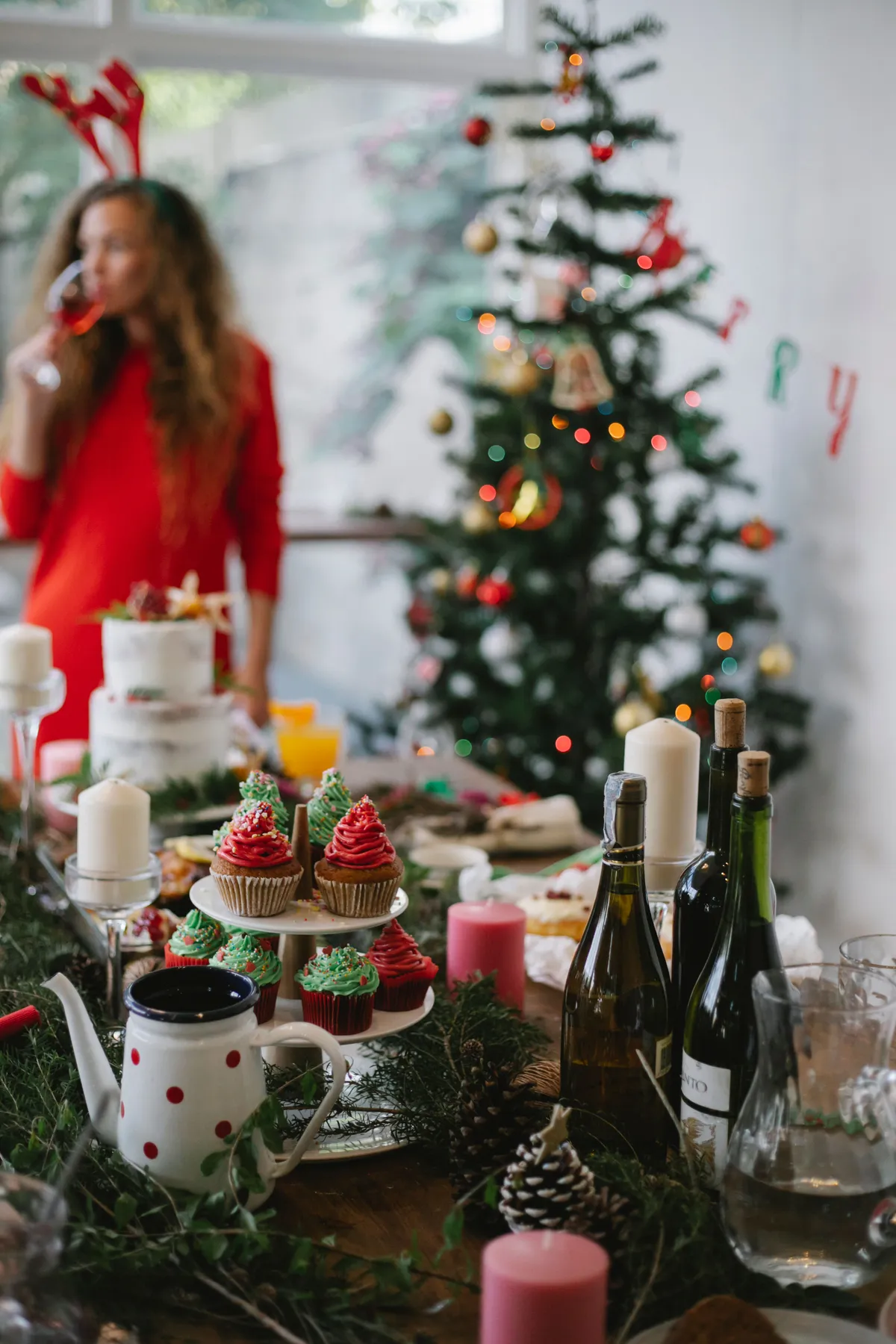 Female in antler headband drinking champagne standing near table with dinner for Christmas celebration and fir tree