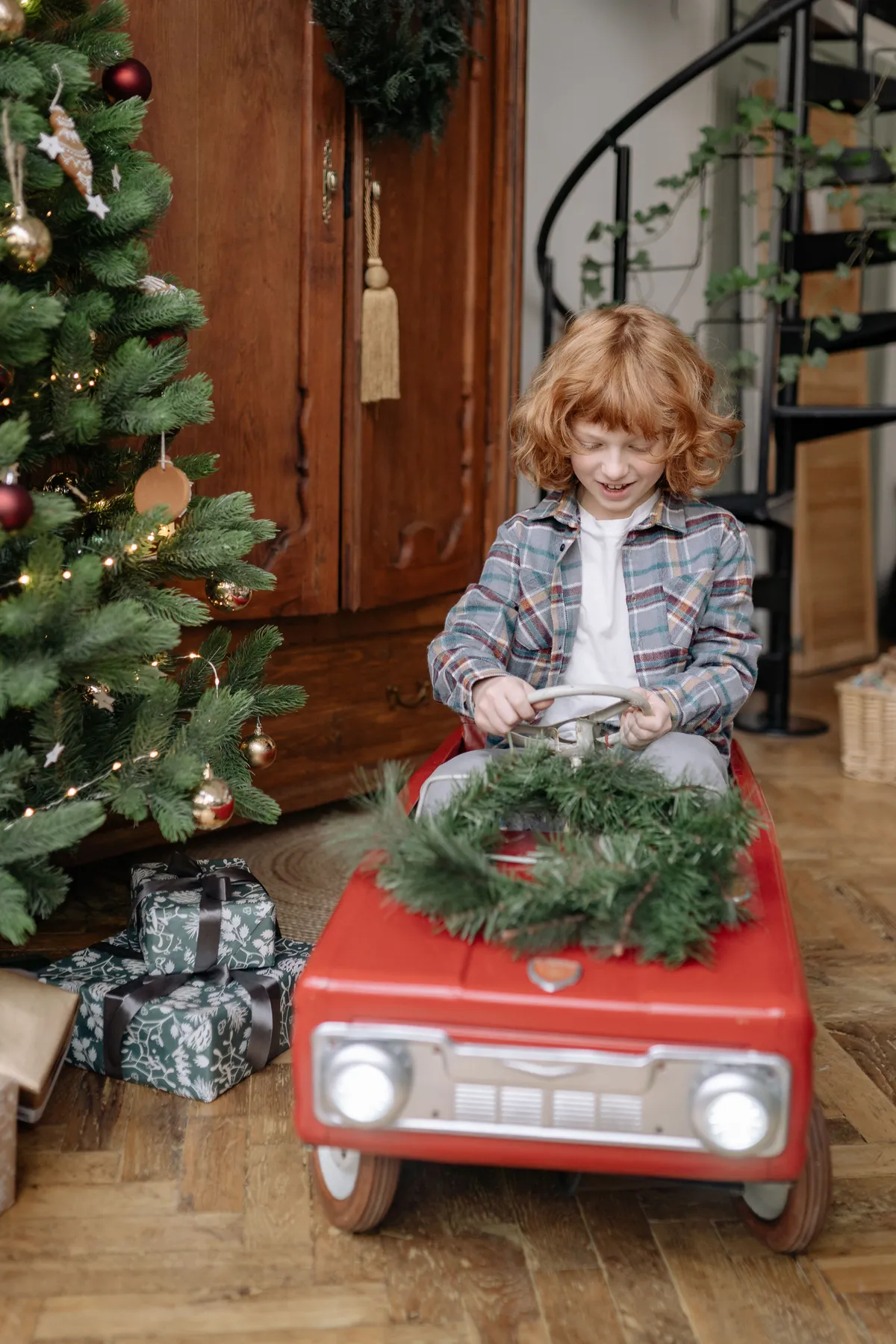 Happy child in toy car beside Christmas tree, enjoying festive indoor celebration.