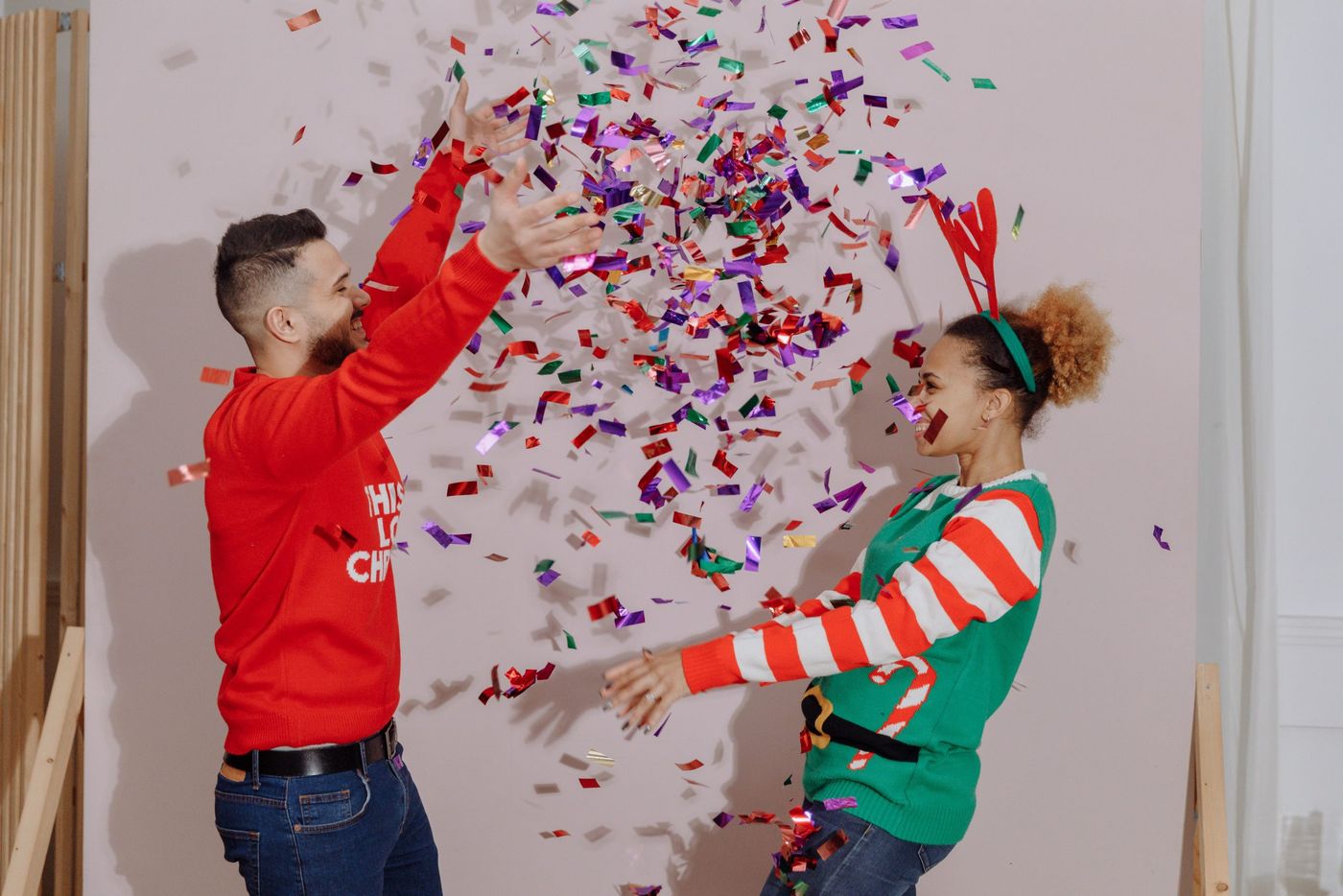 Interracial couple celebrating holidays with colorful confetti in festive sweaters indoors.