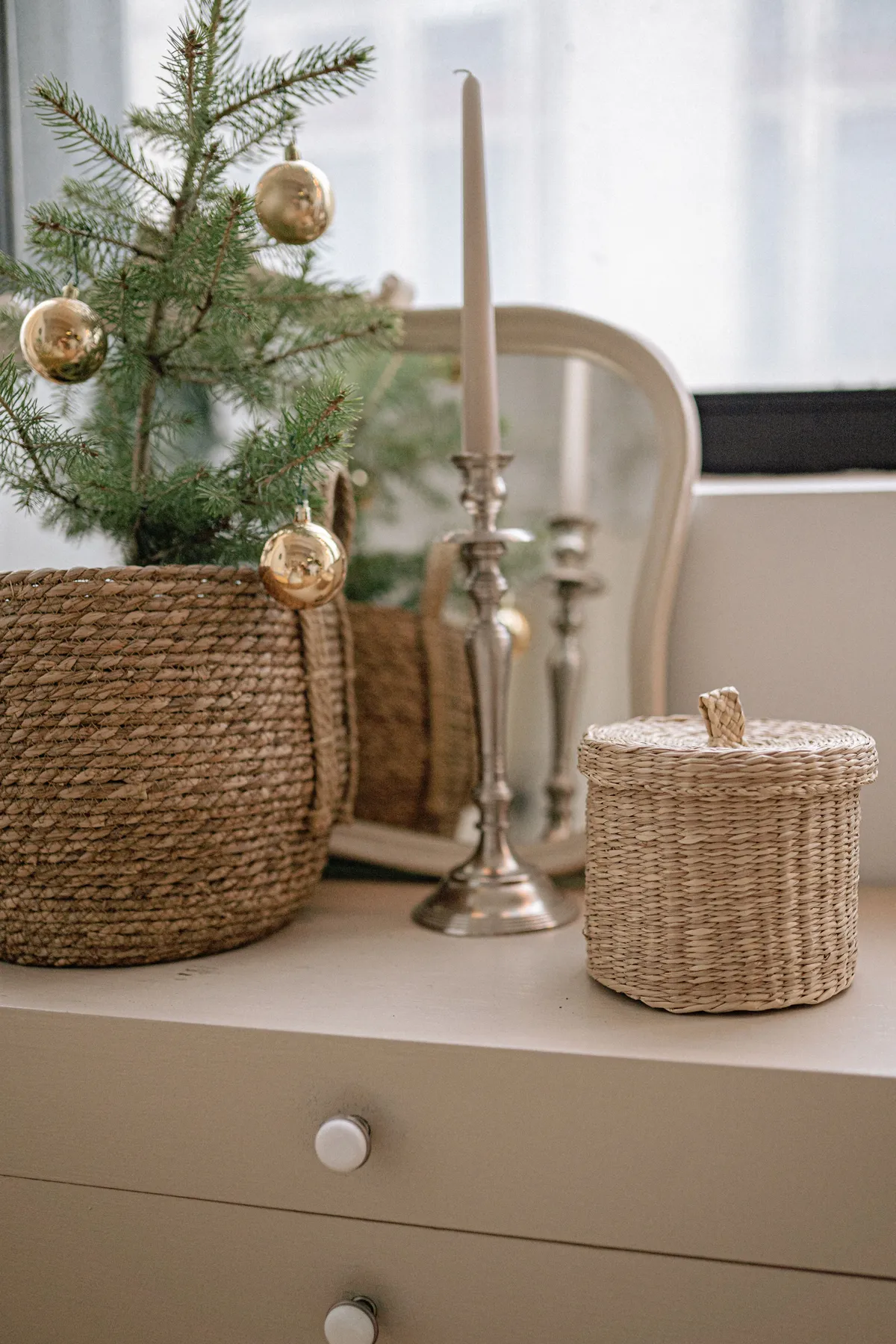 Stylish holiday setup with a candlestick, mirror, and festive pine in a wicker basket on a dresser.