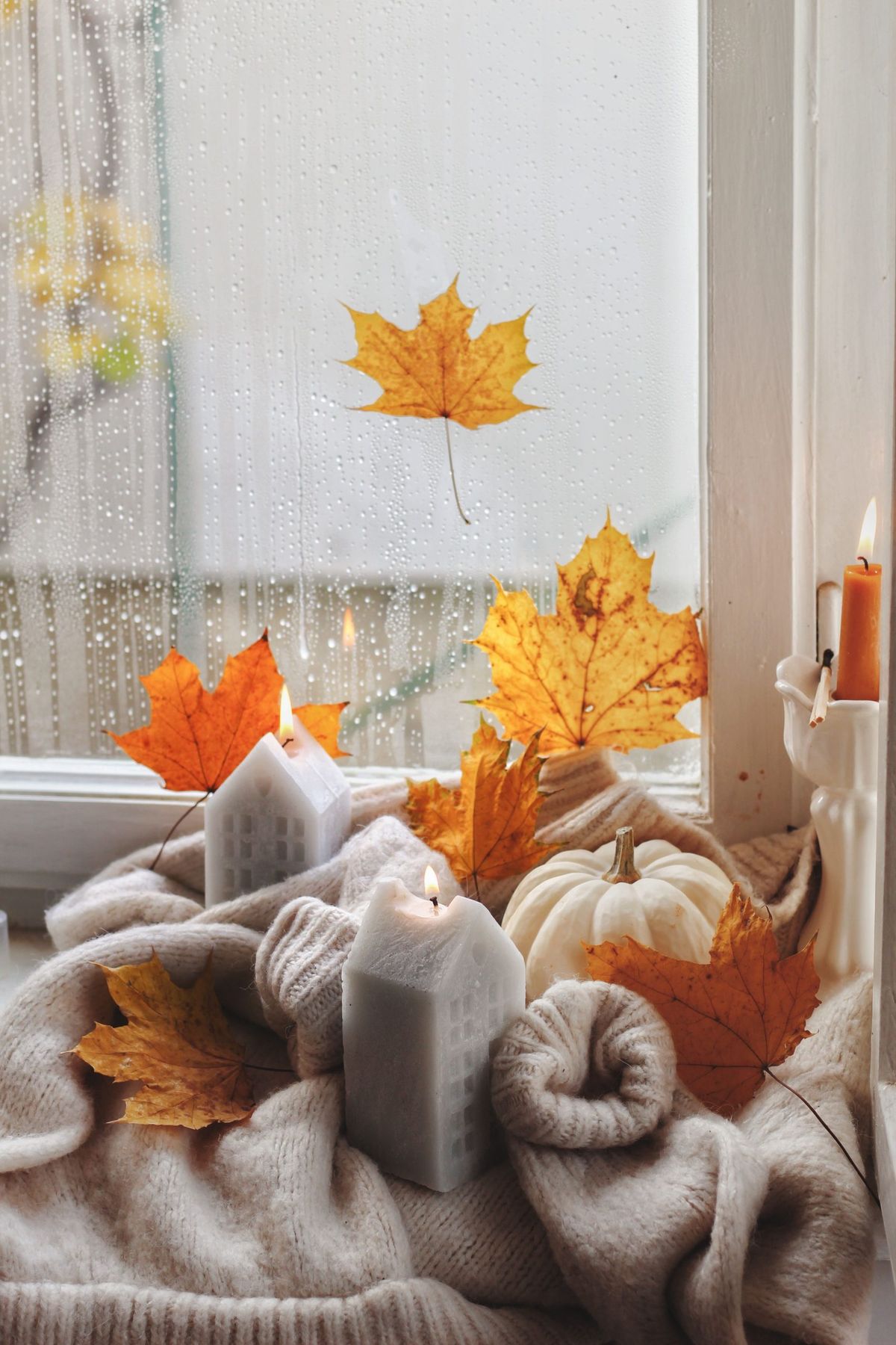 Warm and cozy fall setting featuring candles, a sweater, and autumn leaves by a rainy window.