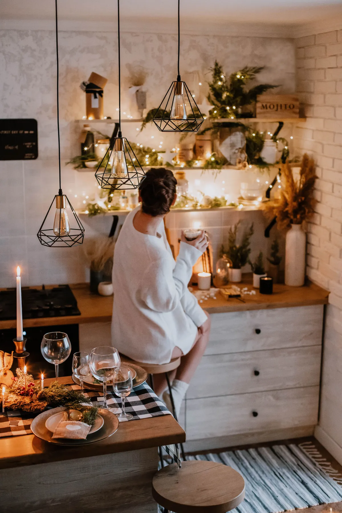 Warm indoor scene of a woman in a cozy kitchen during the holidays, with decorative lights.