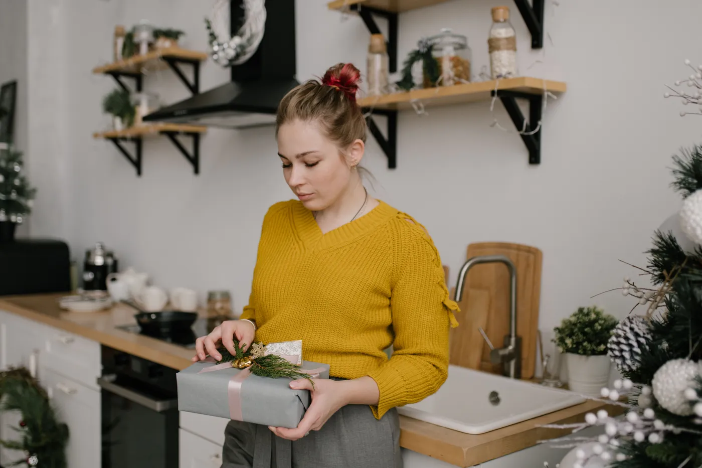 Young woman in yellow sweater holding a gift box in a cozy, decorated kitchen during winter.